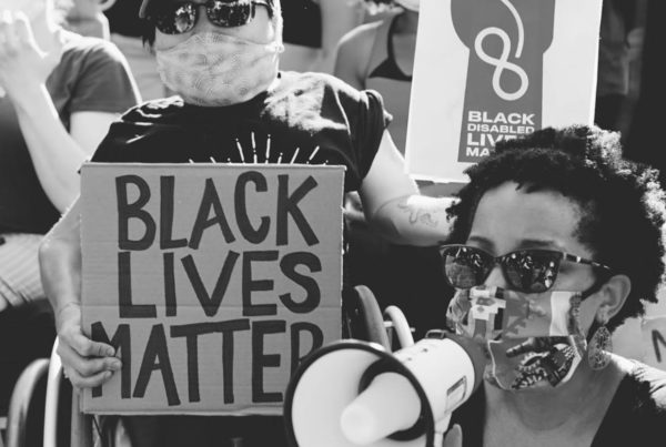 A black and white photo of Keri Gray and Mia Ives Rublee at a protest. Keri is speaking on a bullhorn and Mia is holding signs that say "Black Lives Matter."