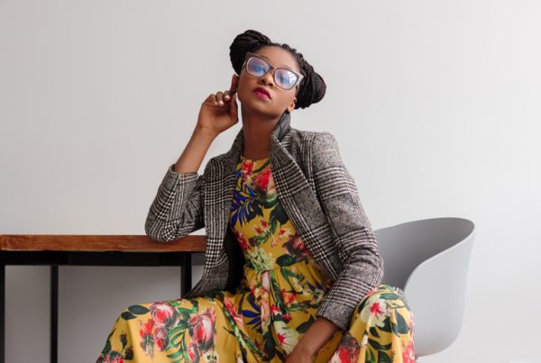Photo of a confident young Black woman. She is posing in a simple office setting. She is wearing a yellow flower dress, gray blazer, glasses, and her braids are in two side buns.
