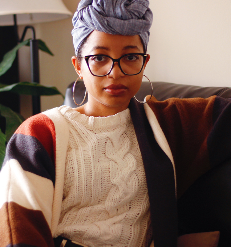 Keri Gray sitting on a couch in a white sweater, multicolored sweater jack and blue jean headwrap.