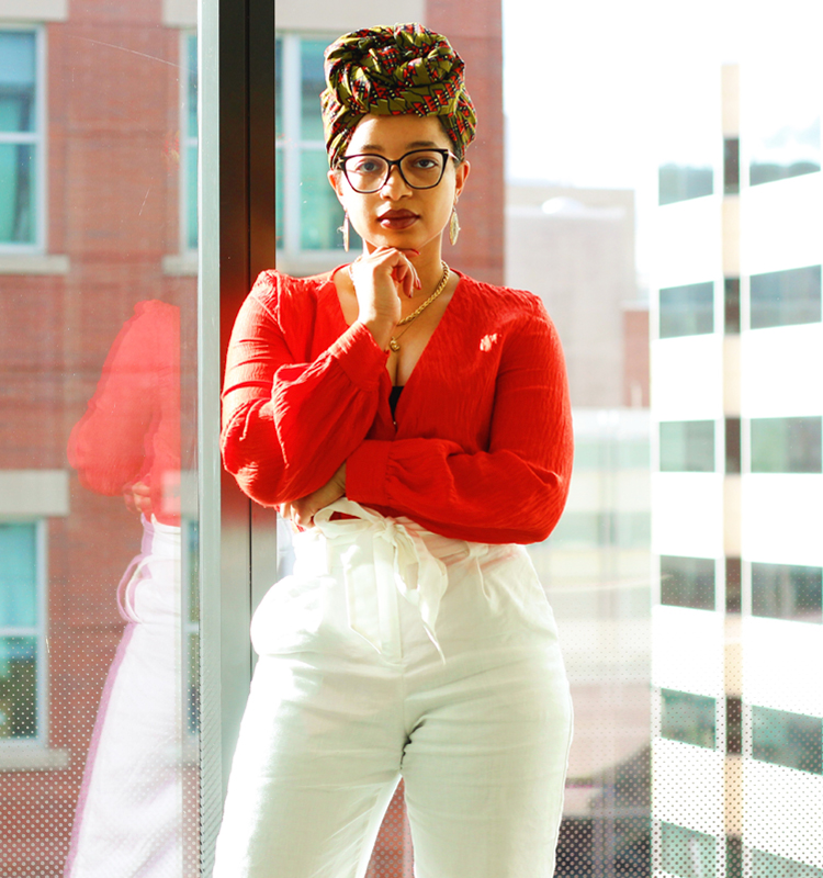 Photo of Keri wearing an orange shirt, white pants, and green and orange headwrap. She appears to be in an office with a large glass window with a couple of building in the background.