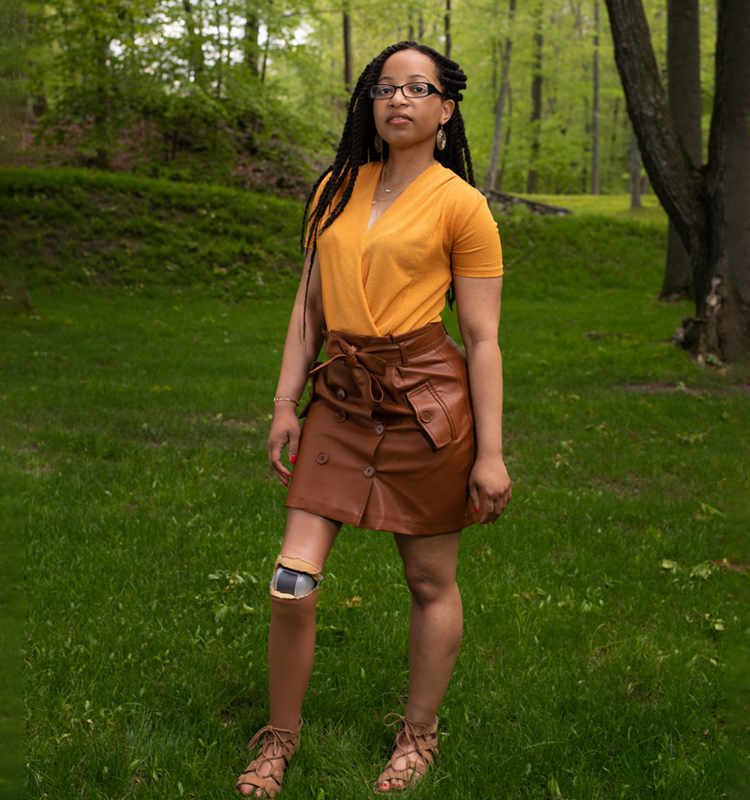 Photo of Keri Gray in a mustard shirt, brown skirt and long twists. She is standing in a very green setting of grass and hills.