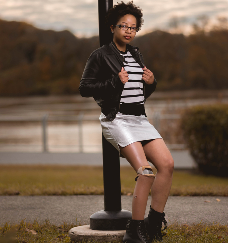 Keri Gray in a silver skirt, black and white shirt, and black jacket. She is posing against a pole with a park/water in the background.