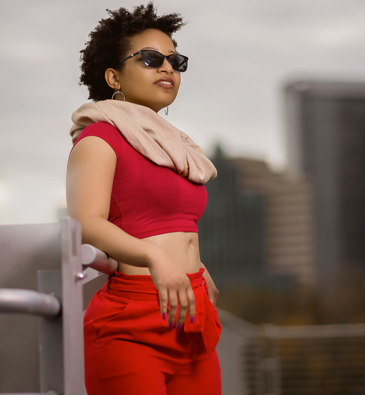 Keri Gray in a red cut off shirt, red pants, sunglasses and a tan scarf. She is standing against a rail and blurred building are in the background.