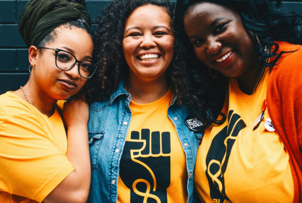 Three Black women are standing in front of a black brick wall, they are each wearing a yellow t-shirt that says ‘Black Disabled Lives Matter’ inside of a Black fist. The top of the photo has white text that says Our Presence is Our Power * November 8, 2021. @namdadvocates