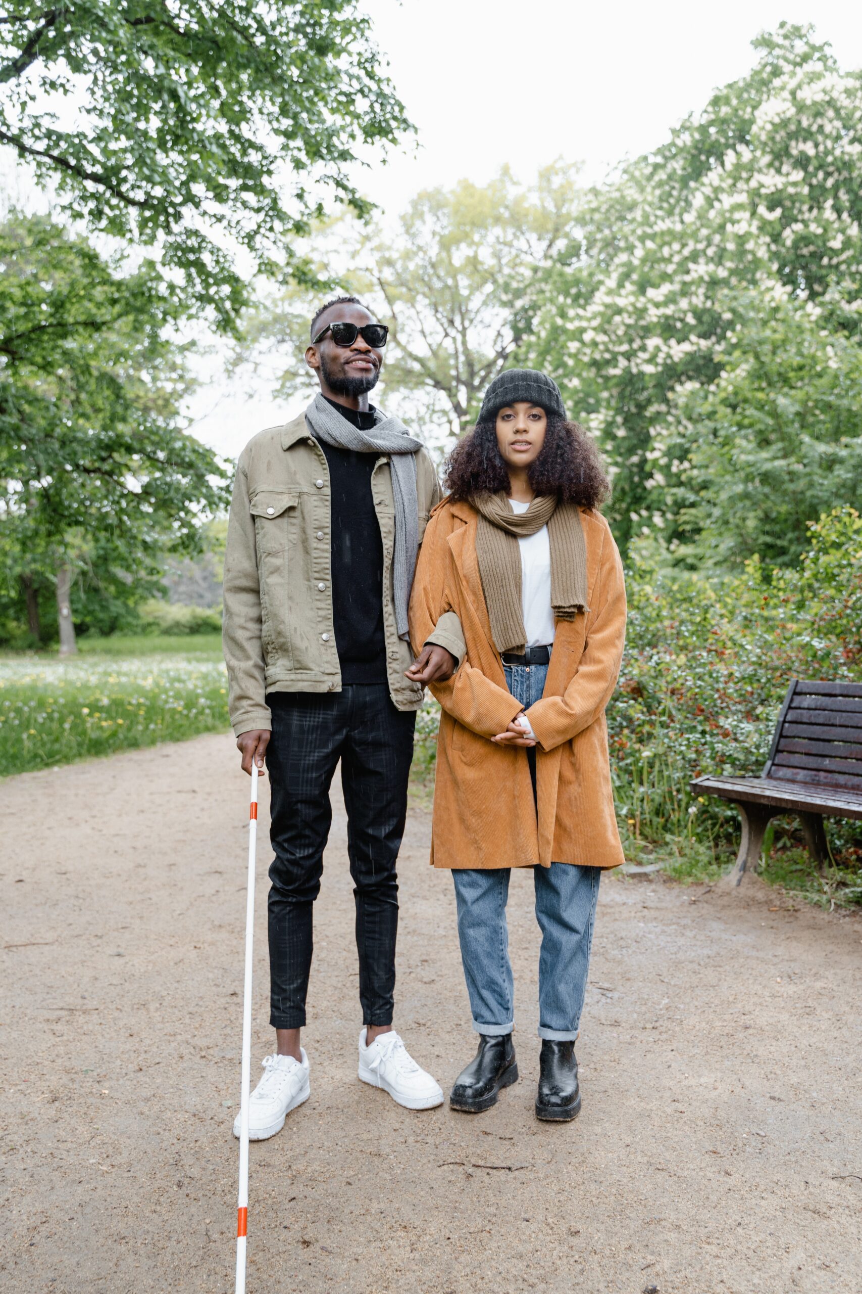 Image of two Black people standing in a park. The male on the left has a white cane.