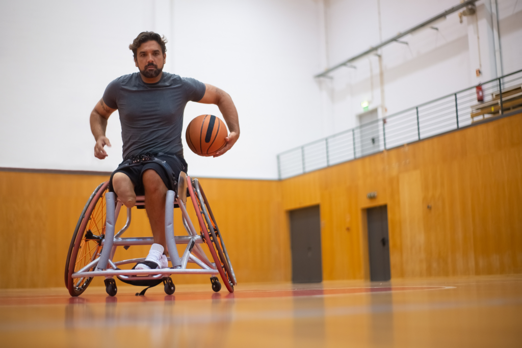 A medium-skinned man with an amputation is in a sports wheelchair at a gymnasium. He's holding a basketball in one hand.