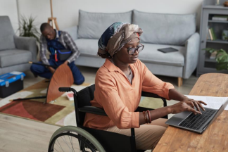 At a desk, a dark-skinned woman in a wheelchair is using her laptop. She is in a cozy living room.