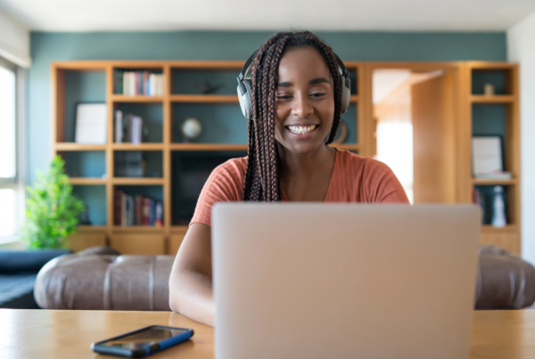 At home, a young woman with dark skin and long, brown locs is working on her laptop while wearing headphones. She's smiling.
