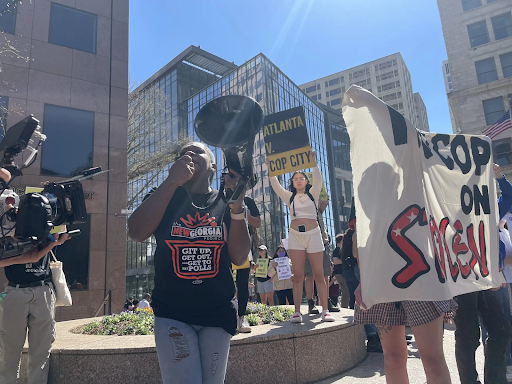 A dark-skinned individual holding a megaphone, addresses a crowd of Stop Cop City protesters in front of the Georgia Pacific Building in downtown Atlanta.
