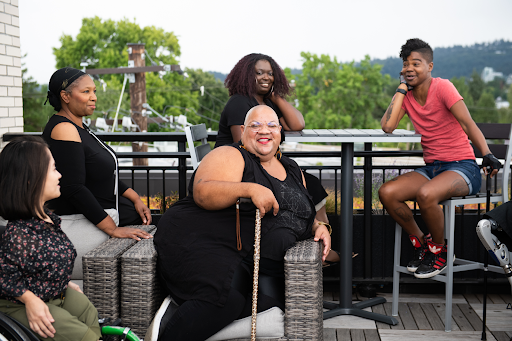 A Black non-binary person laughs while sitting in a cushioned wicker chair with their leopard print cane. They are surrounded by four more disabled people of color. Everyone is in the midst of conversation at a rooftop deck party.