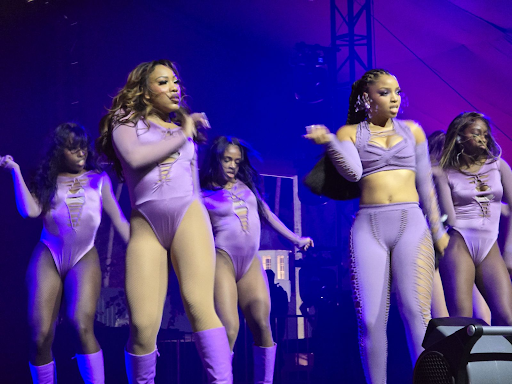 A group of femme black backup dancers and a performer are dancing on the stage at the Coachella Music Festival. They are all wearing purple attire and striking fierce poses. The background is bathed in purple lighting, and a large black speaker is positioned at the front of the stage.